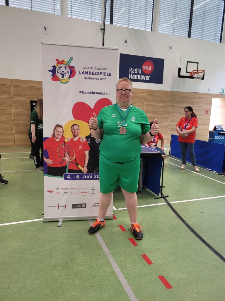 Foto von Anika Huskamp, die in Sportkleidung in einer Turnhalle steht. Sie trägt eine Bronze-Medaille und zeigt mit beiden Händen Daumen hoch.