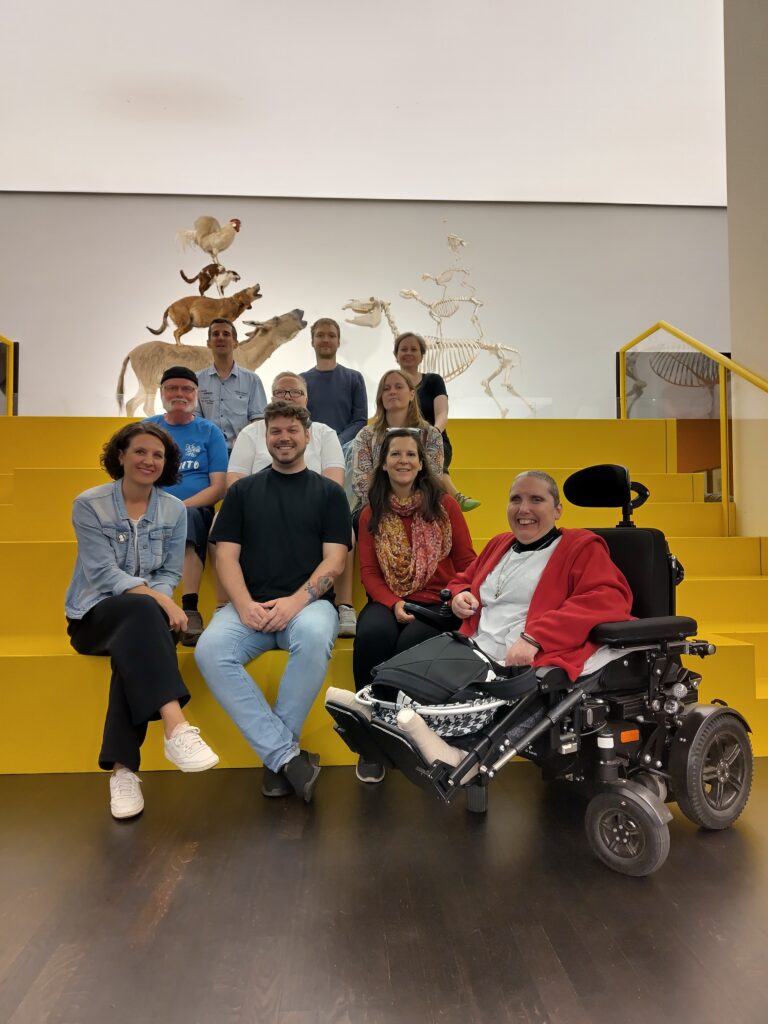 Alle Kollegen aus dem Büro auf einem Foto. Wir sitzen auf einer großen gelben Treppe. Hinter uns sind Kunstwerke von den Bremer Stadtmusikanten.