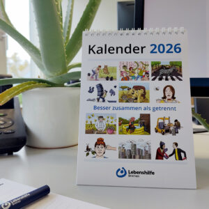 Foto von dem Kalender 2026 auf einem Schreibtisch. Man sieht am Rand vom Bild zum Beispiel einen Block mit Stift, Telefon, Pflanze und Bildschirm.